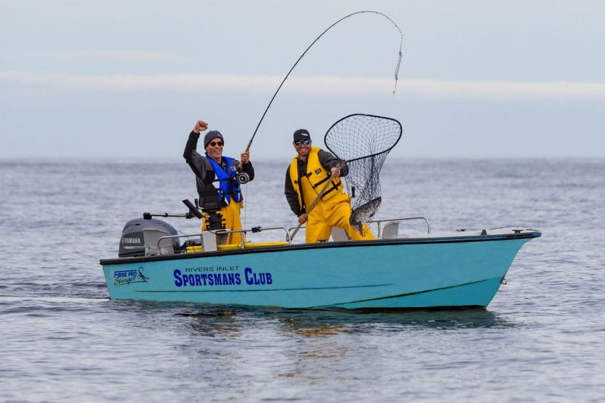experience-2021-mod Two men in a Rivers Inlet Sportsman's Club boat in the open ocean. One man is holding the net and in it is a big BC salmon. The other man is holding the fishing rod and raising his fist in the air celebrating their accomplishment. They look thrilled on their fishing trip in BC, Canada.
